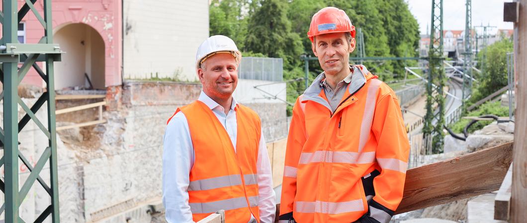 Bagger fahren im Gleisbereich, ein Krahn befördert Baumaterial von der einen Brückenseite auf die andere und Arbeiter stemmen Beton von den Bohrpfählen.